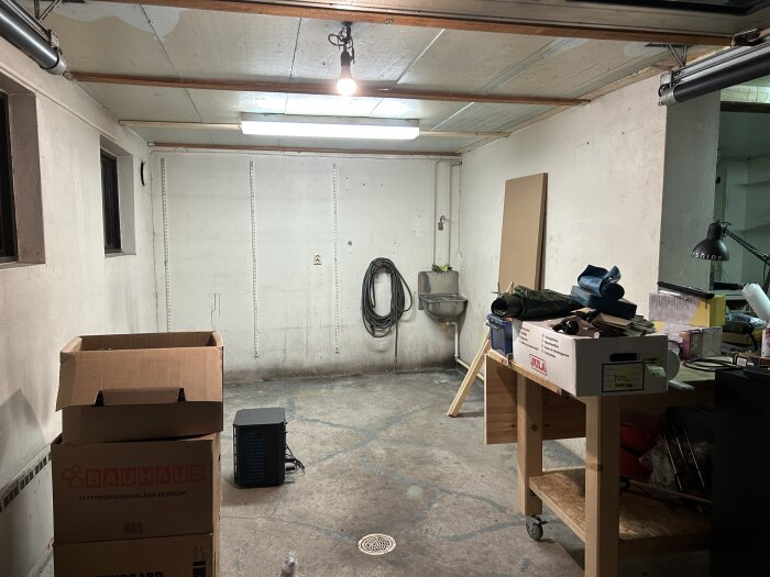 Dubbelgarage med betonggolv, kartonger, verktyg på bord och en väggmonterad diskho. Takbelysning och kablar längs väggarna.