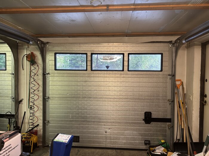 Dubbla garageportar i metall med fönster, omgivna av några verktyg och trädgårdsredskap längs väggarna i ett oinrett garage.