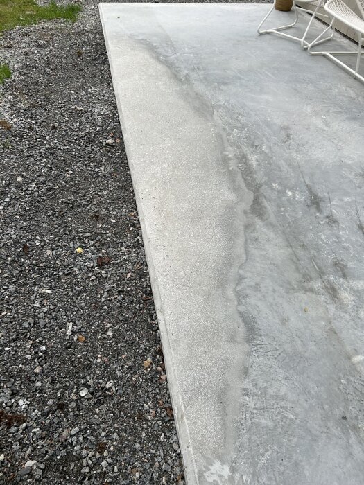 Betongplatta med ojämn yta visar framträdande ballast och skålformade områden, omgiven av grus och två stolar i bakgrunden.