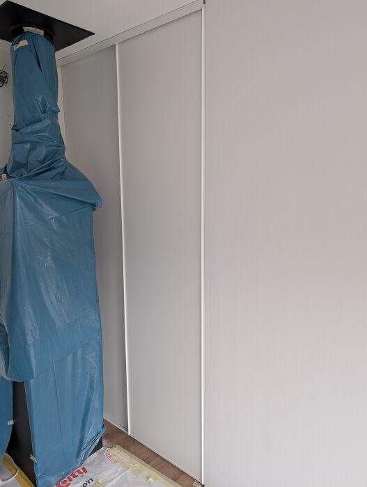 Skjutdörr monterad mot innervägg med synligt glapp. Plast skyddar en del av väggen intill, trägolv och målat gipstak syns.