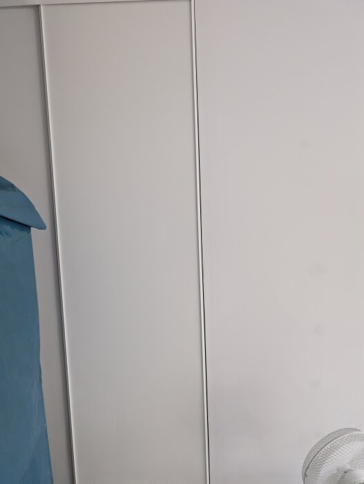 Skjutdörr monterad mot en vit gipsvägg med glapp mellan dörr och vägg, visar ojämt hörn. Fläkt och täckt möbel syns delvis.