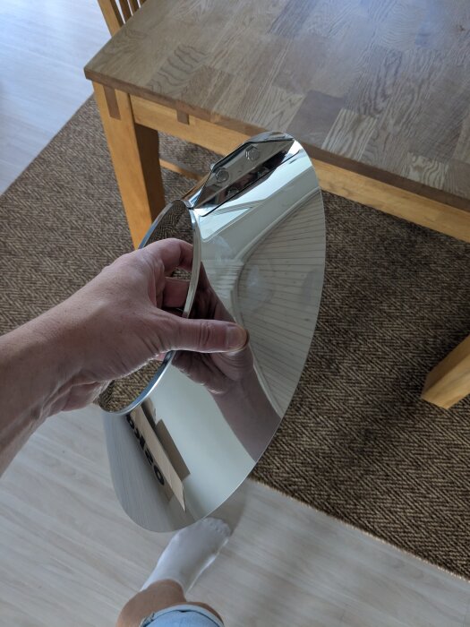 En person håller en blänkande metallisk regnkrage för skorsten framför ett träbord.