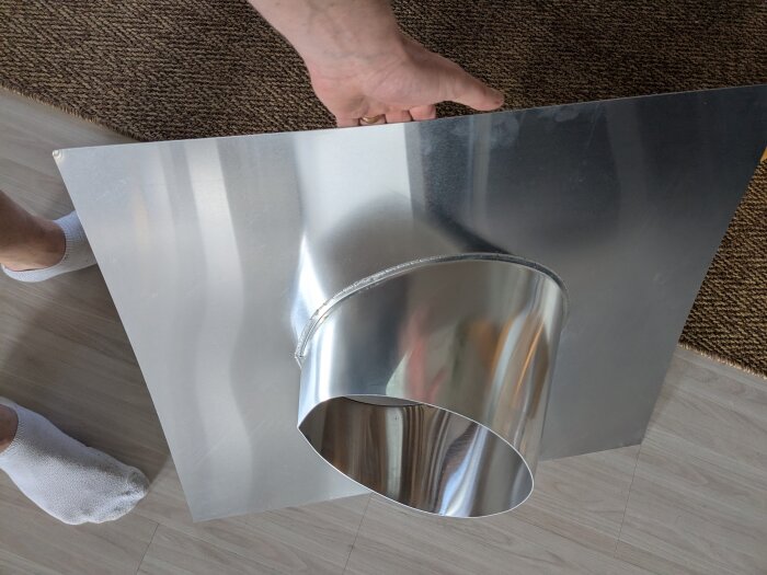 Metallplåt för skorsten med hål, hålls av en hand inomhus. Synlig fot med vit strumpa på golv.