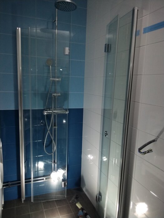 Duschområde i badrum med blå och vit kaklad fondvägg, glasdörrar och arbetsbelysning. Blå tejp syns på en av väggarna.
