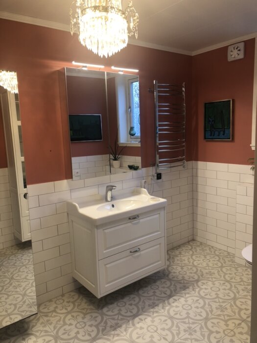 Modernt badrum med vit kommod, mönstrat golv, spegel med LED-belysning, handdukstork och vägg i tegelstensmönster under blandande färger.