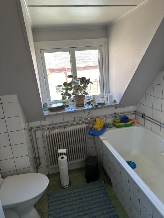 Litet badrum med vita kaklade väggar, badkar, toalett, radiator och växter i fönster på hylla. Barnleksaker placerade på badkarskanten.