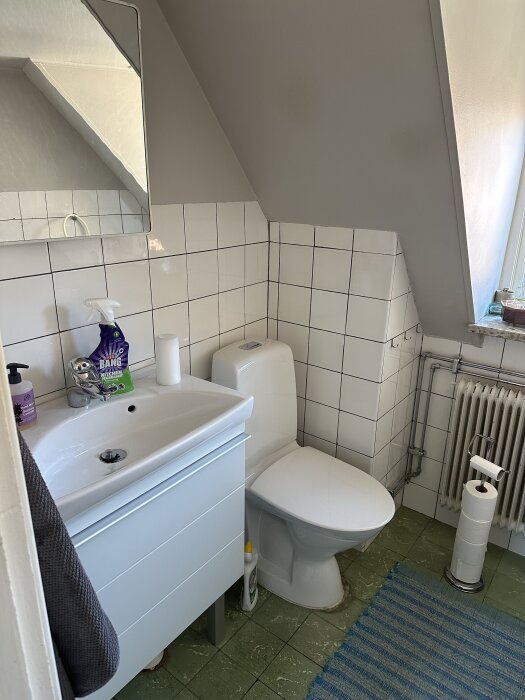 Litet badrum med kaklade väggar, grönmelerat klinkergolv, vit toalett, handfat med spegel samt element under snedtak.