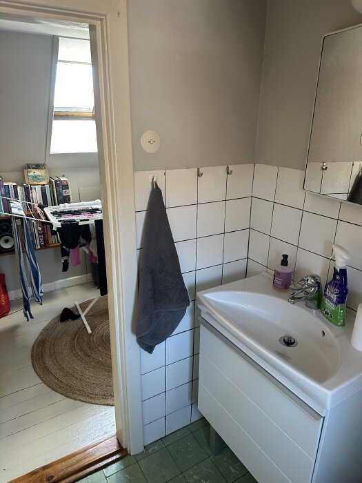 Litet badrum med handfat, spegel och vit kakelvägg, synlig dörröppning mot tvättområde med hyllor och kläder.