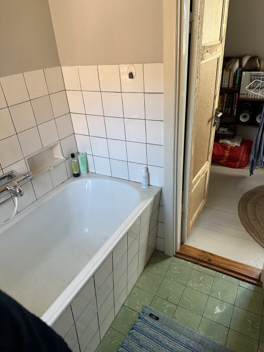 Gammalt badrum med vitt kakel och grönt golv, äldre badkar, del av dörr och entré skymtar. Toalettartiklar syns på badkarskanten.