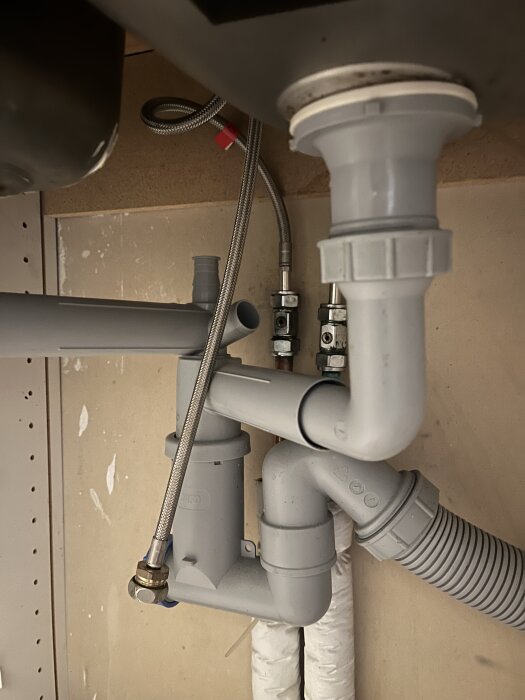 Utrymmet under diskhon visar rörsystem med gråa avloppsrör och metallslang, potentiellt för en bänkdiskmaskin.