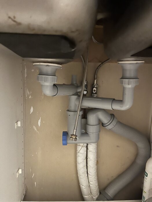 Rörsystem under diskbänk med anslutningar för vatten och avlopp, inklusive grå avloppsslang och vitisolerade rör, redo för bänkdiskmaskin.