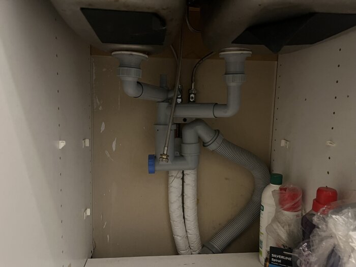 Diskbänksskåp under diskho med rör och avloppsanslutningar synliga; plats för att installera en kompakt diskmaskin.