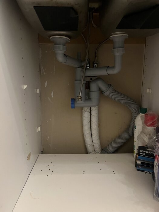 Utrymmet under diskhon med synligt avloppsrör och vattenanslutningar. En flaska rengöringsmedel och andra hushållsartiklar vid sidan.