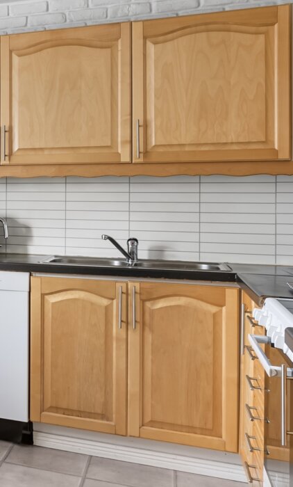 Kök med ljusbruna skåpluckor, silverfärgade handtag, rostfri diskbänk och vitkaklad vägg.