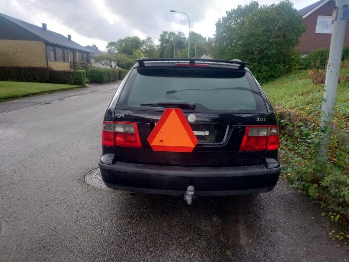 Svart bil med en nybyggd hållare för en orange LGF-skylt på bakluckan, fotograferad på en gata med hus och träd i bakgrunden.