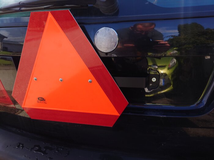 Orange LGF-skylt monterad på svart bakre fordonspanel, spegling av person i bakgrunden.