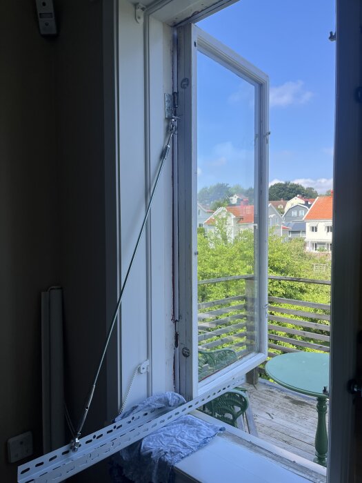 Hemgjord fönsterlyft med hyllkonsoler och gängstång håller upp ett öppet fönster mot en balkong med utsikt över ett grönområde och hus.