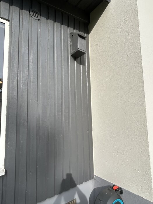 Uterumspanel målad i Jotum Aska, med grundfärg i Jotun Shadow. Solbelyst eftermiddagsscen med skugga av fotografen och en rulle för slang synlig.