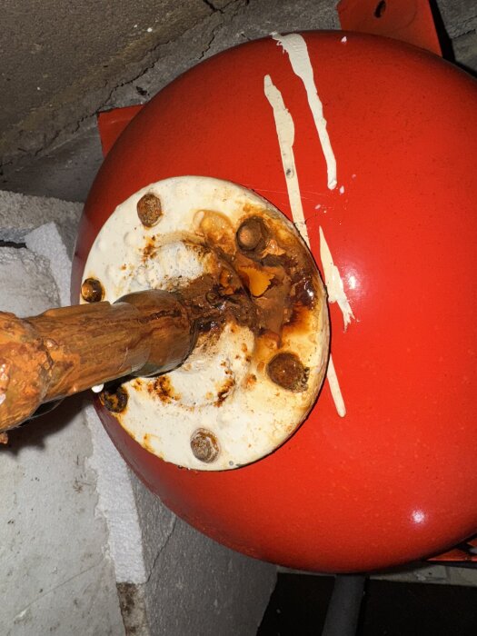 Rostig hydropress med vattenläcka i en krypgrund, röd yta med vita etiketter, en kopparröranslutning synlig.