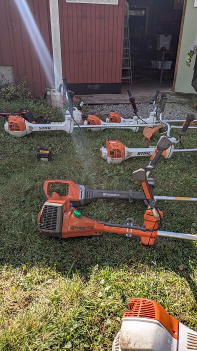 Flera grästrimmers av märkena Stihl och Husqvarna står på en gräsmatta framför en röd byggnad med en öppen dörr.