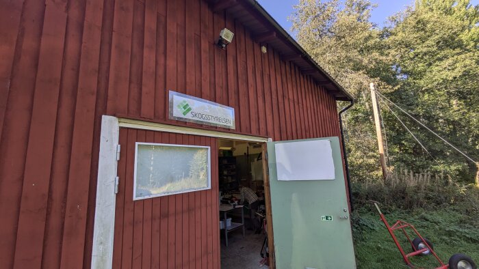 Röd byggnad med öppen dörr, skylt med texten "Skogsstyrelsen" ovanför. Grönt gräs och en röd skottkärra i förgrunden, skog i bakgrunden.