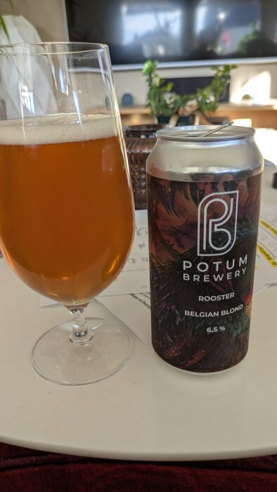 Ölglas och burk med Potum Brewery Rooster Belgian Blond, 6,5%, på ett bord inomhus.