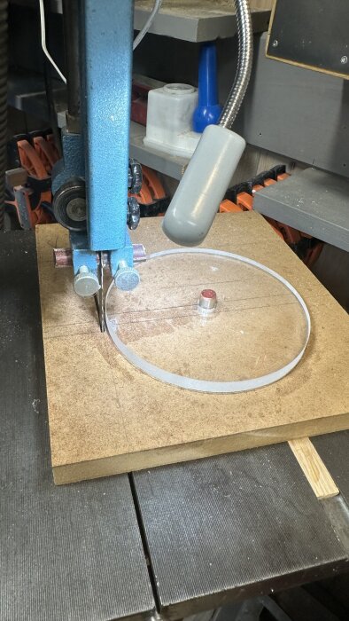 Bandsåg skär en cirkel av plexiglas, omgiven av verktyg och klämmor på ett arbetsbord.