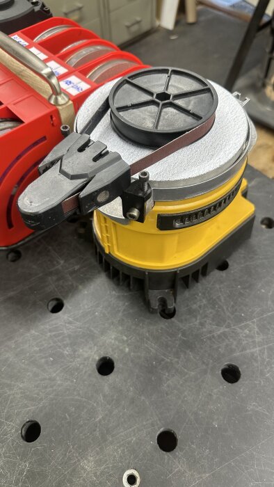 Slipmaskin Work Sharp WS2000 med slipband och justerbar jig för verktyg, placerad på en arbetsbänk. Röd fackförvaring i bakgrunden.