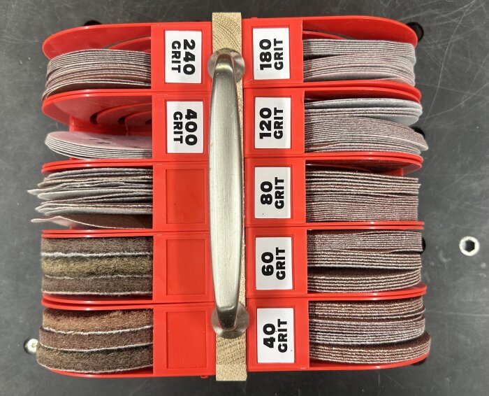Förvaring med slipmaterial i olika grovlekar (40-400 grit) i röd plasthållare med handtag, synliga etiketter och skivor i olika lager.