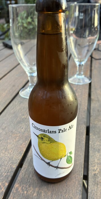 Flaska med närproducerad Citronärlans Pale Ale står på ett träbord med två tomma glas i bakgrunden.