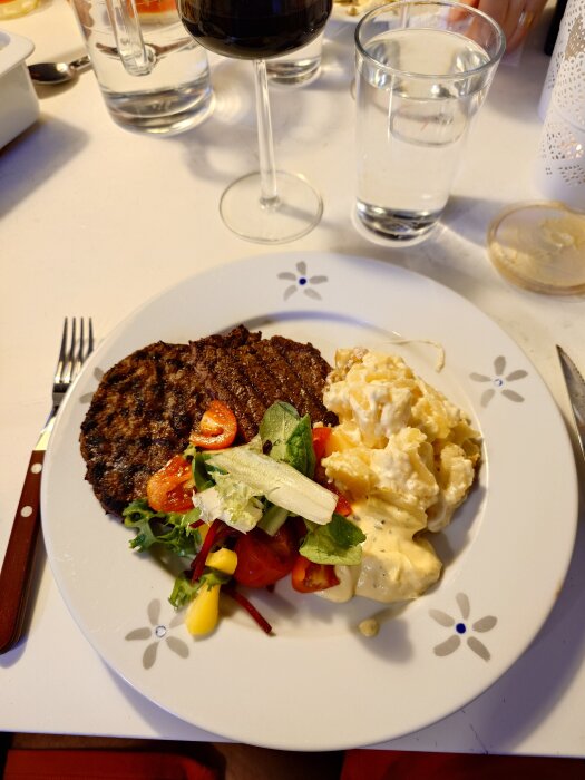 Tallrik med stödburgare, sallad, potatissallad och sås, omgiven av glas vatten och vin på ett dukat bord.