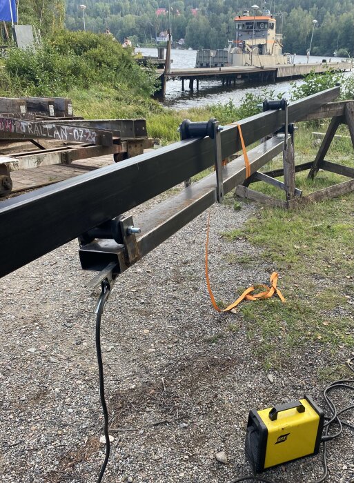Ett svartmålat, svetsat metallrör monterat på rullar med orange rem, vid en hamn med båt i bakgrunden. Gul svetsutrustning syns på marken.