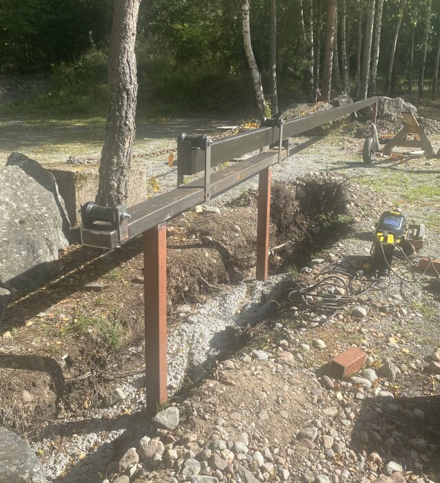 Svetsad metallbom på rullar, placerad utomhus vid grusunderlag. Området omges av träd och stenar, och svetsutrustning syns på marken.
