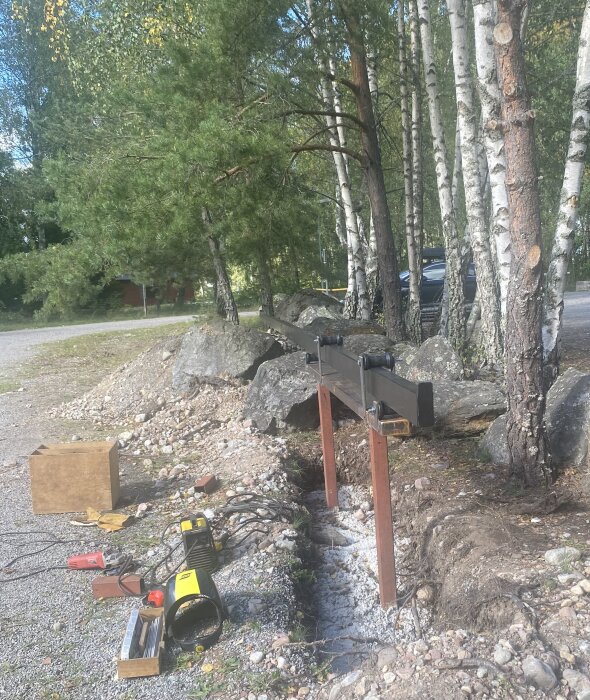Svart stålrör monterat på två röda metallstolpar i en grusad väg, omgiven av träd och stenar; svetsutrustning i förgrunden.