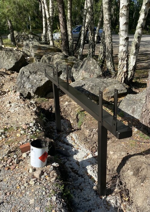 Svartmålad metallbom monterad mellan stolpar i skogsmiljö, omgiven av stenar och träd. Färgburk syns i förgrunden.