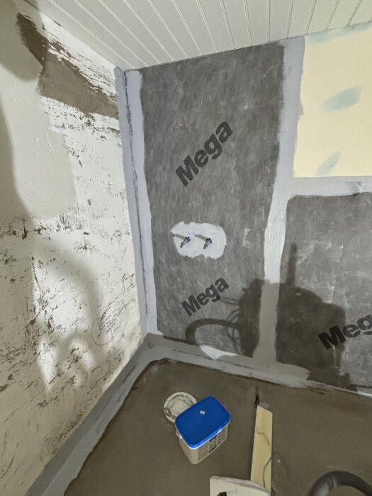 Vägg med isoleringsmaterial märkt "Mega", cementgolv, plastburk med blått lock och verktyg i ett hörn under renovering.