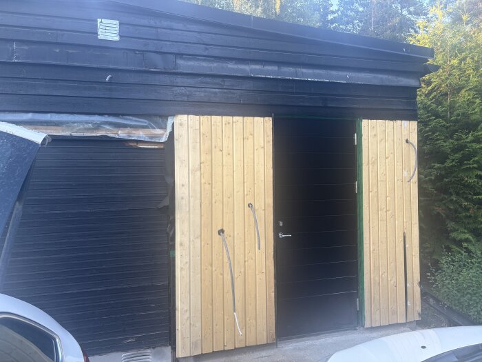 Svart garage med fönsterlös dörr, träpaneler på sidorna och platt tak, omgivna av grönska och en del av en bil synlig i förgrunden.