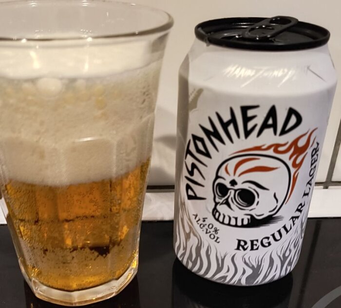 En burk Pistonhead Regular Lager 4% och ett glas öl med skum på toppen står på en bänk.