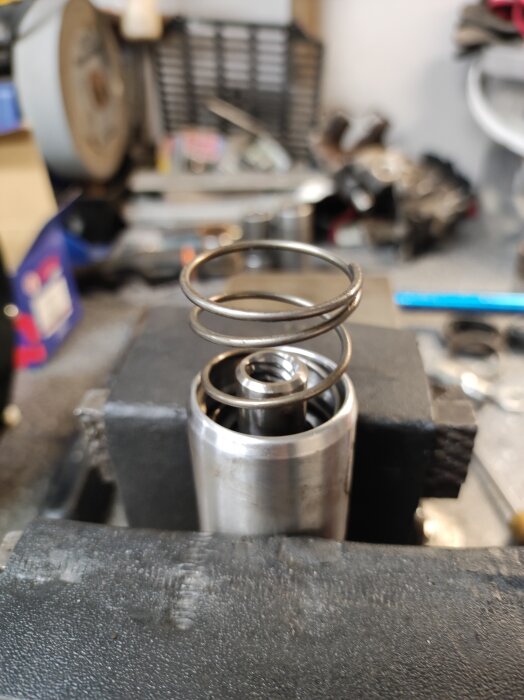 Metallfjäder i en cylinder i ett verkstadsutrymme, omgiven av verktyg och föremål i bakgrunden.