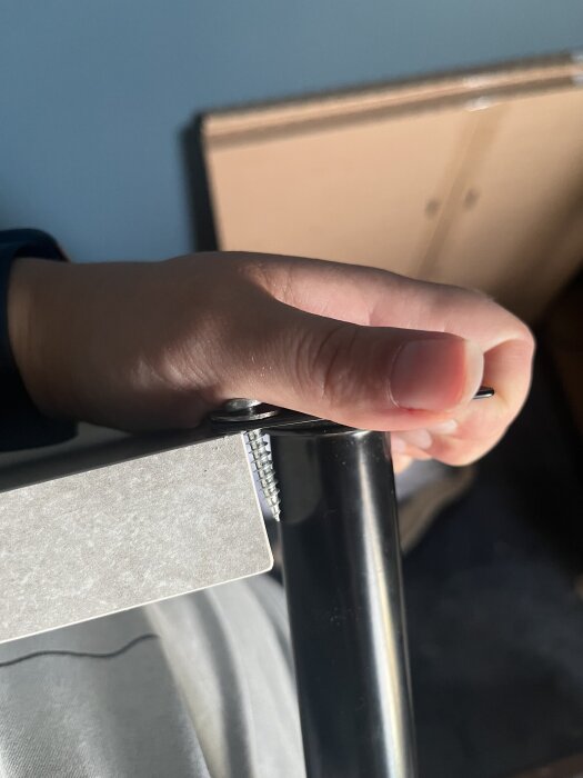 En hand håller en skruv mot en laminatskiva och ett metallben. Skruven verkar för lång för skivan.