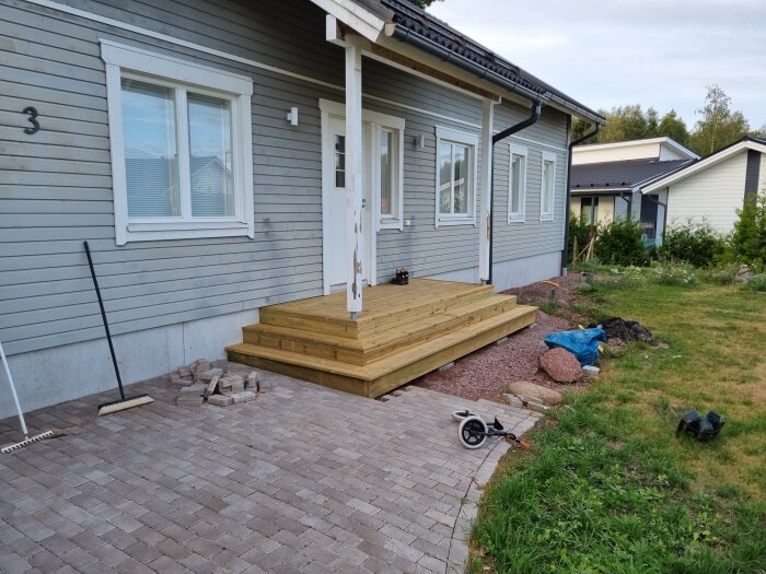 Ny stenlagd yta framför grått trähus med trätrappa. Krattor och stenar syns på marken. Gräsmatta delvis synlig.