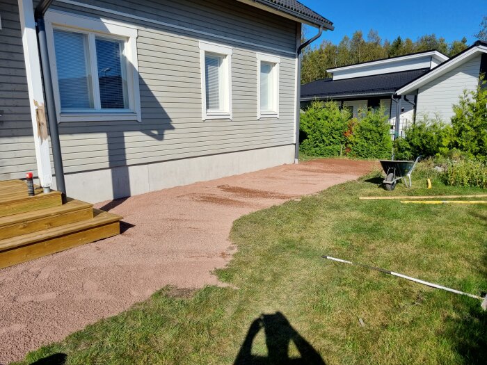 Markberedning för stenläggning med utlagt grus intill ett hus, skottkärra och verktyg syns på gräsmattan.