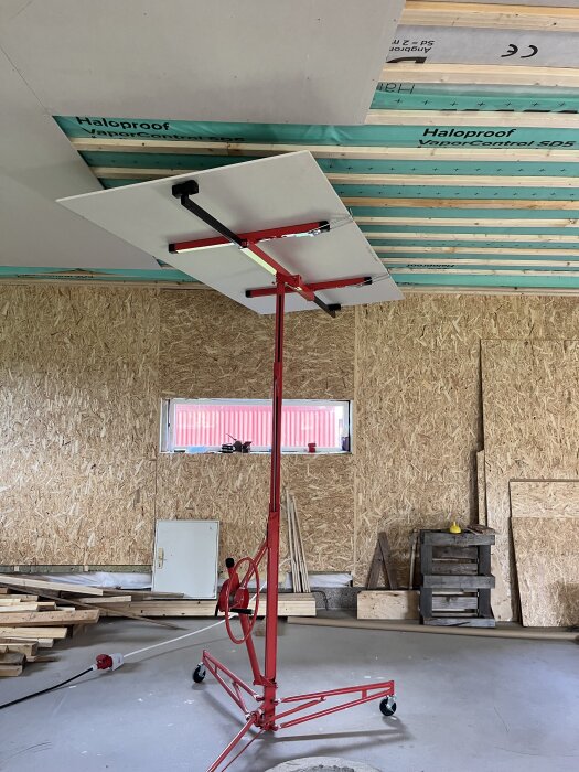 En gipsskiva monteras i taket med hjälp av en röd skivlyft i ett rum under renovering.