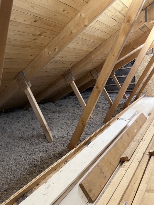Innertak med träbjälkar och isolering på vinden, redo för gipsning. Hyllor och plankor ligger staplade på golvet.