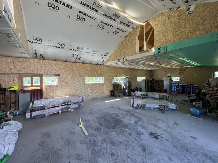 Byggarbetsplats med isolerat tak och material staplat på golvet, innertak med plastmaterial, samt verktyg och utrustning runt om.