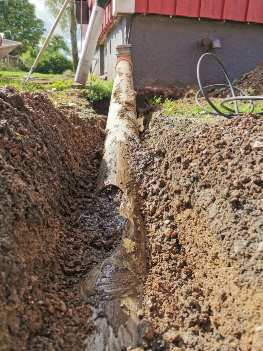 Trasigt dagvattenrör nära husgrund i en grävd ränna med jord runtom, solsken ovan.