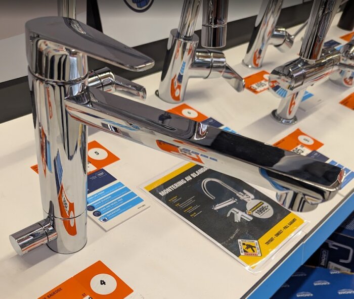 Kromad köksblandare med inbyggt handtag ovanpå, placerad i en butiksmiljö på en vit yta med broschyrer och prisetiketter.