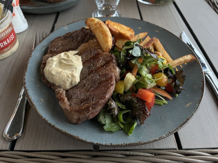 Tallrik med grillad entrecote, bearnaisesås, potatisklyftor och sallad med grönsaker på ett bord.