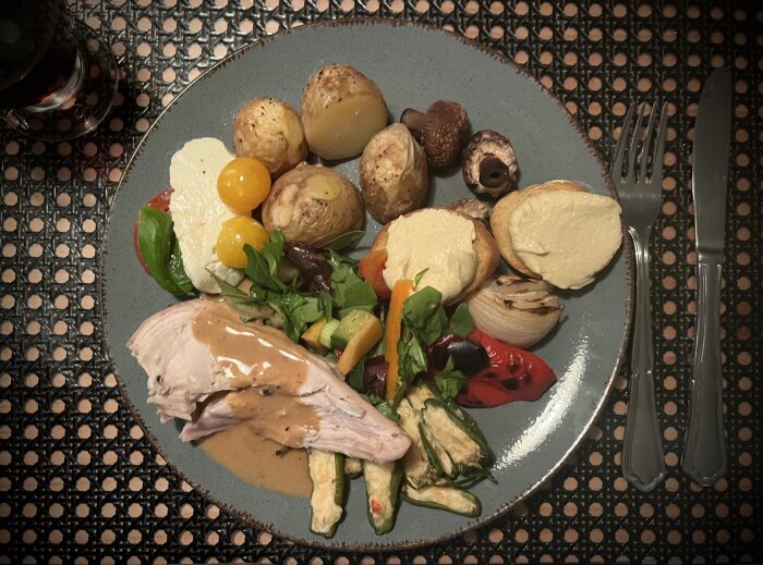 Tallrik med grillad kyckling, ugnstekt potatis, crostini med hummus, caprese och grönsaker, serverad med sås och dryck vid sidan.