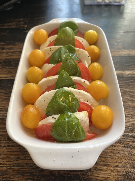 Caprese-sallad i en vit form med tomater, mozzarella, basilika och gula cocktailtomater på ett mörkt träbord.